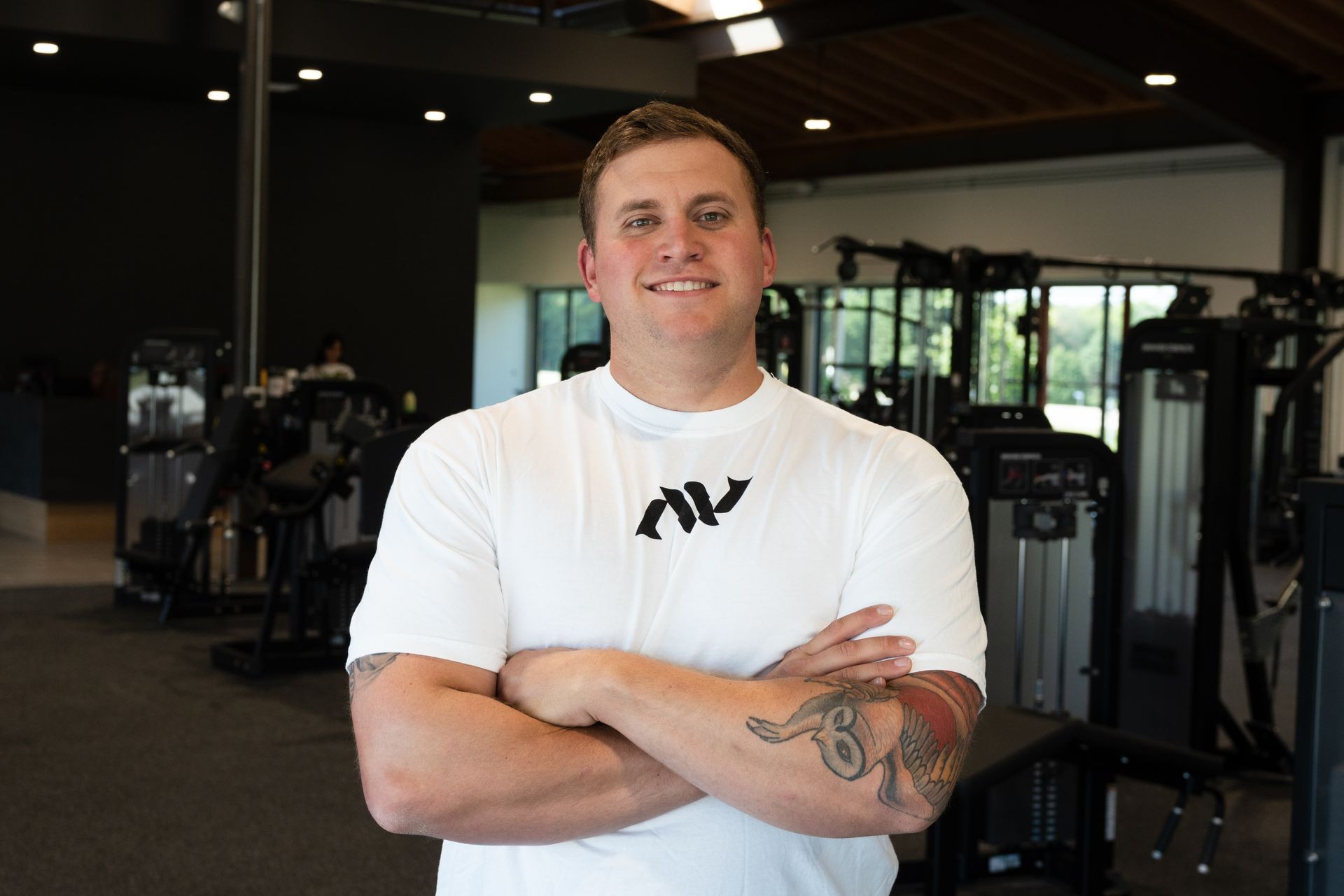 Person with a tattoo on their arm standing in a gym with various fitness equipment in the background.