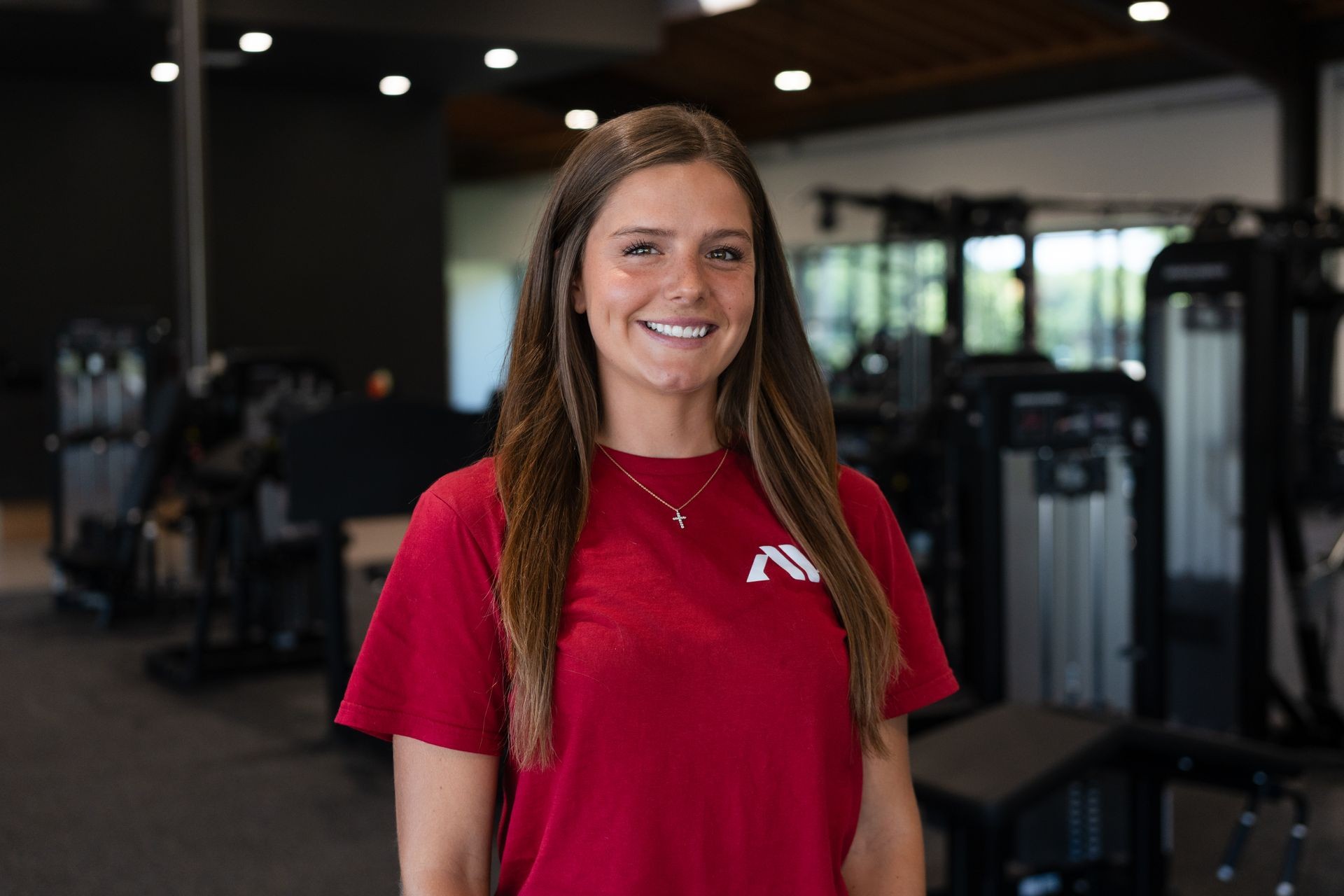 Woman in a red shirt standing and smiling in a gym with exercise equipment in the background.