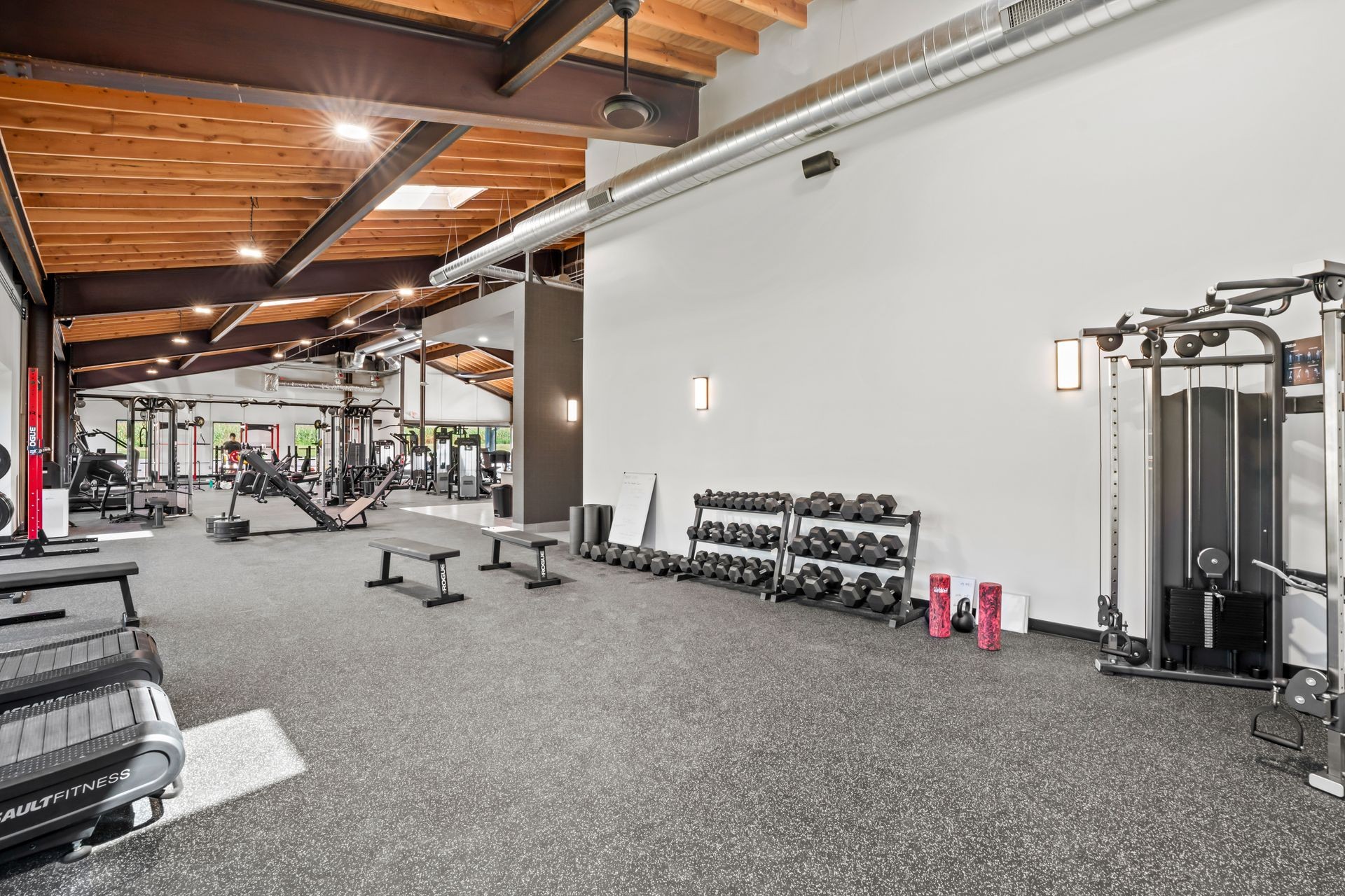 Modern gym interior with various exercise equipment, weights, and benches under an open wooden ceiling.