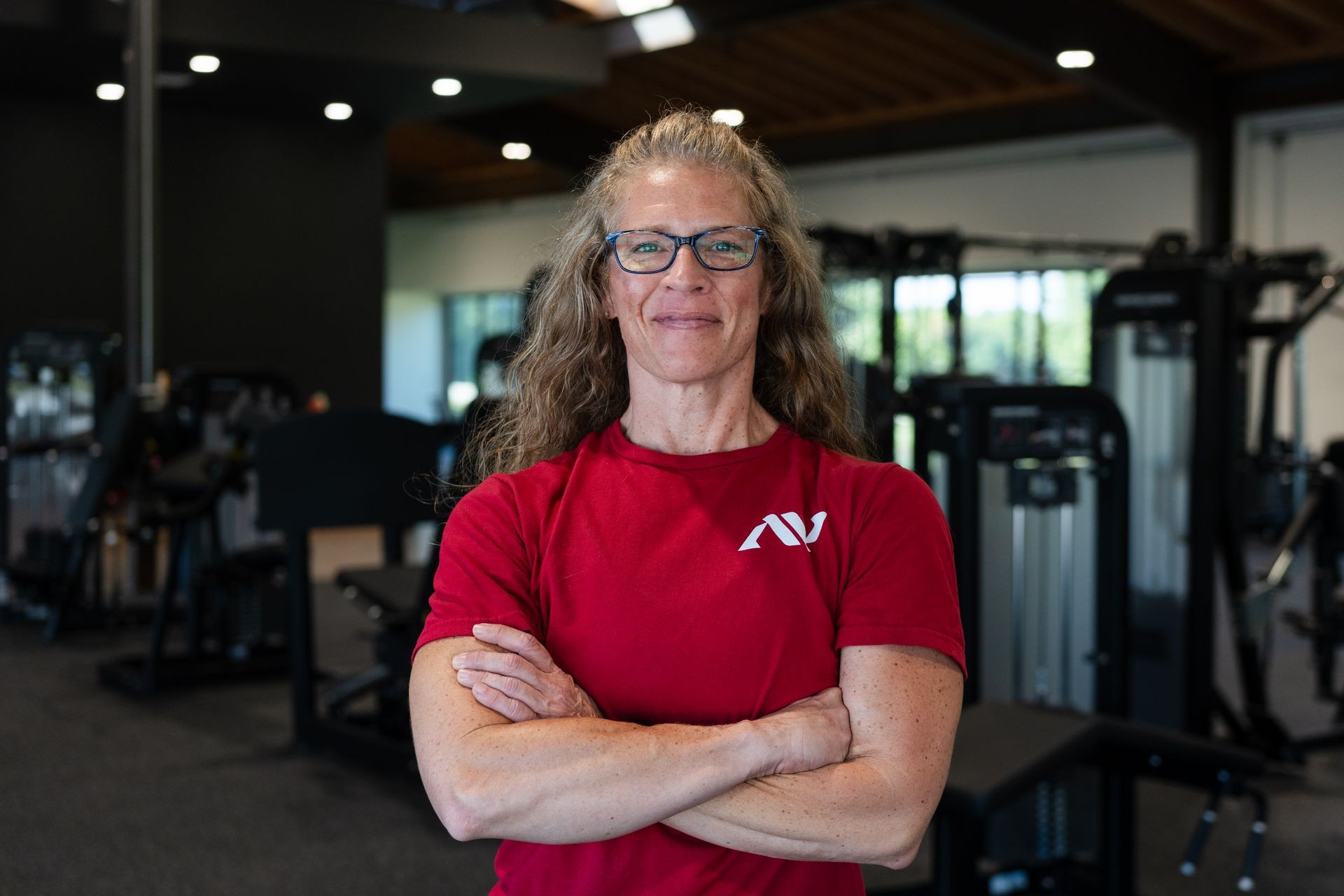 Person in a red shirt with crossed arms standing in a gym, surrounded by various exercise equipment.